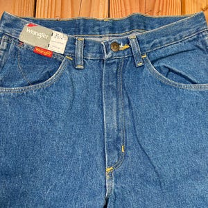 May include: Close-up of blue denim Wrangler jeans with a button closure and yellow stitching. The jeans have front and back pockets and a Wrangler tag. The waist size is 31 inches and the inseam is 32 inches.