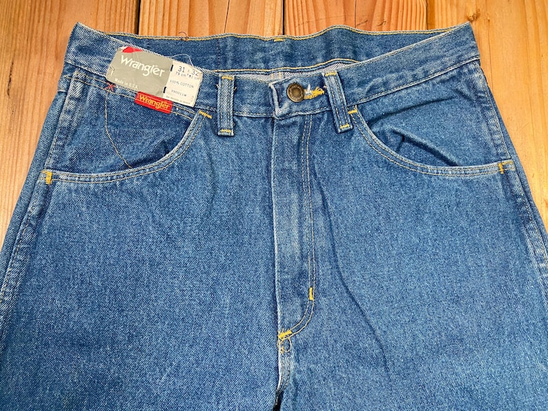 May include: Close-up of blue denim Wrangler jeans with a button closure and yellow stitching. The jeans have front and back pockets and a Wrangler tag. The waist size is 31 inches and the inseam is 32 inches.