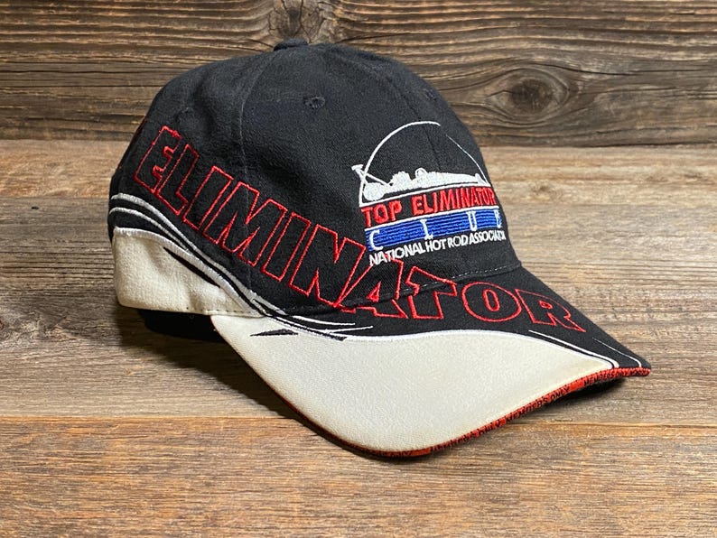 May include: Black baseball cap with "ELIMINATOR" in red lettering. The cap features a white bill with red trim and the text "TOP ELIMINATOR CLUB NATIONAL HOT ROD ASSOCIATION". The hat is likely a collectible item.