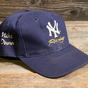 May include: A navy blue baseball cap featuring an embroidered white New York Yankees logo on the front with the word Racing in gold script below it. The side of the cap displays the embroidered text Nike Dunn in gold script lettering.