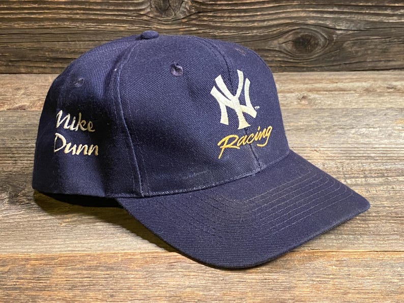 May include: A navy blue baseball cap featuring an embroidered white New York Yankees logo on the front with the word Racing in gold script below it. The side of the cap displays the embroidered text Nike Dunn in gold script lettering.