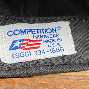 May include: Close-up of a black baseball cap with a white tag. The tag features the text "COMPETITION HEADWEAR MADE IN U.S.A." and a blue star and red stripes graphic.