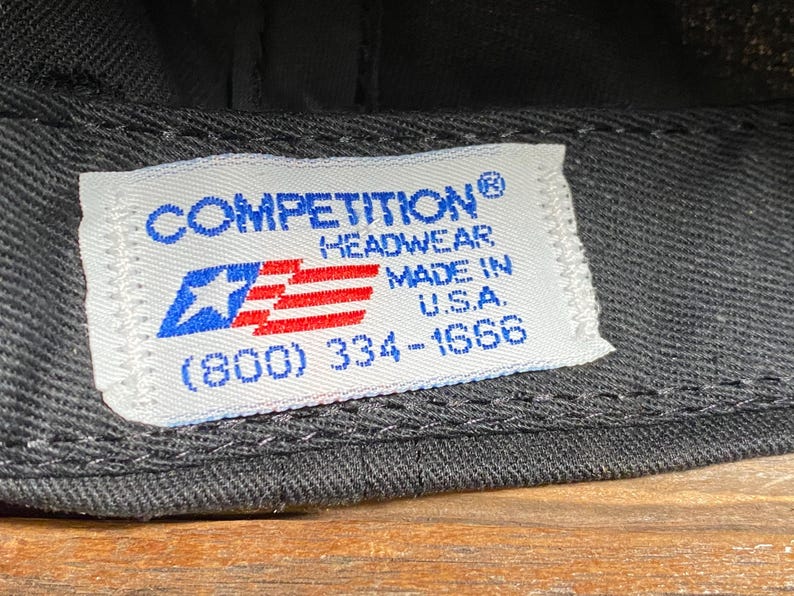 May include: Close-up of a black baseball cap with a white tag. The tag features the text "COMPETITION HEADWEAR MADE IN U.S.A." and a blue star and red stripes graphic.