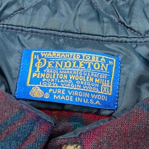 May include: Close-up of a blue Pendleton label stitched onto a quilted, dark teal fabric. The label reads "WARRANTED TO BE A PENDLETON" and includes text about the wool and origin. The garment is a red and blue plaid.