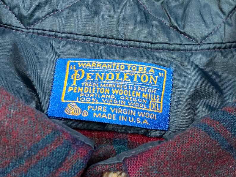May include: Close-up of a blue Pendleton label stitched onto a quilted, dark teal fabric. The label reads "WARRANTED TO BE A PENDLETON" and includes text about the wool and origin. The garment is a red and blue plaid.