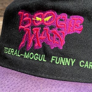 May include: Black baseball cap with a purple brim. The cap features the embroidered text "BOOGIE MAN" in pink with a yellow lightning bolt, and "FEDERAL-MOGUL FUNNY CAR" in green.