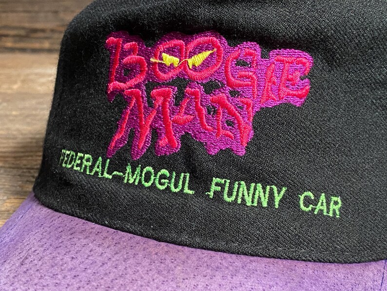 May include: Black baseball cap with a purple brim. The cap features the embroidered text "BOOGIE MAN" in pink with a yellow lightning bolt, and "FEDERAL-MOGUL FUNNY CAR" in green.