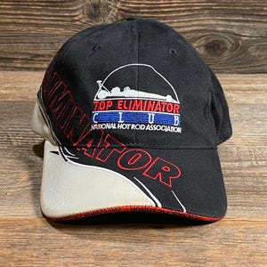 May include: Black baseball cap with "TOP ELIMINATOR CLUB" and "NATIONAL HOT ROD ASSOCIATION" embroidered on the front. The brim is white with red trim. The word "ELIMINATOR" is embroidered in red on the side.