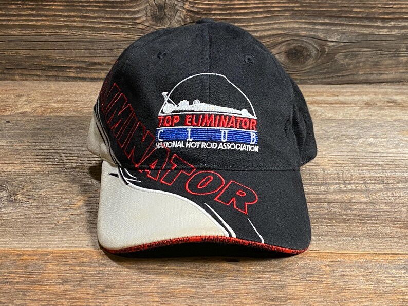 May include: Black baseball cap with "TOP ELIMINATOR CLUB" and "NATIONAL HOT ROD ASSOCIATION" embroidered on the front. The brim is white with red trim. The word "ELIMINATOR" is embroidered in red on the side.