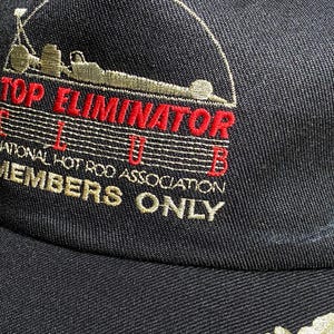 May include: Black baseball cap with embroidered text and graphics. The cap features the words "TOP ELIMINATOR CLUB" in red and "MEMBERS ONLY" in gold. The cap also has a graphic of a race car.