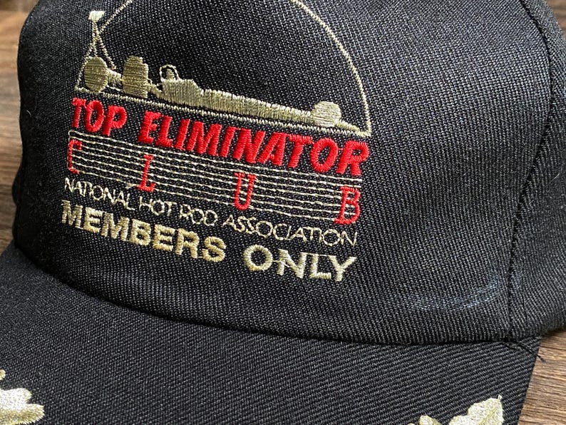 May include: Black baseball cap with embroidered text and graphics. The cap features the words "TOP ELIMINATOR CLUB" in red and "MEMBERS ONLY" in gold. The cap also has a graphic of a race car.