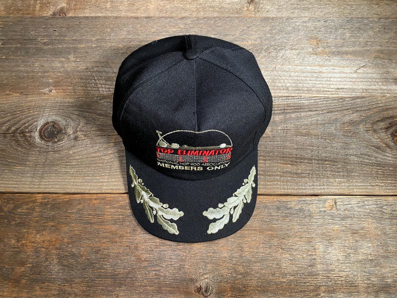 May include: A black baseball cap with embroidered text and design. The text reads "TOP ELIMINATOR NATIONAL HOT ROD ASSOCIATION MEMBERS ONLY". The brim features embroidered green and white leaf designs.