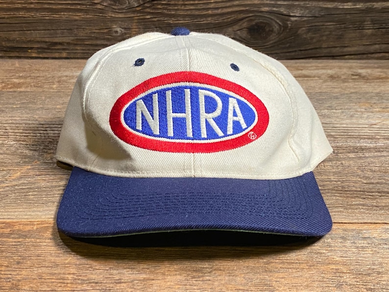 Op de afbeelding: Een witte baseballcap met een marineblauwe klep. De cap toont de letters "NHRA" in blauw, binnen een rode ovaal, op een witte achtergrond. De cap heeft een marineblauwe knop bovenop.