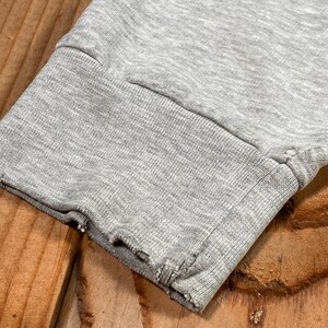 Puede incluir: Primer plano del pu&ntilde;o de una manga de sudadera gris. El pu&ntilde;o es acanalado y tiene un borde deshilachado y desgastado. La tela es gris jaspeado y la manga descansa sobre una superficie de madera.