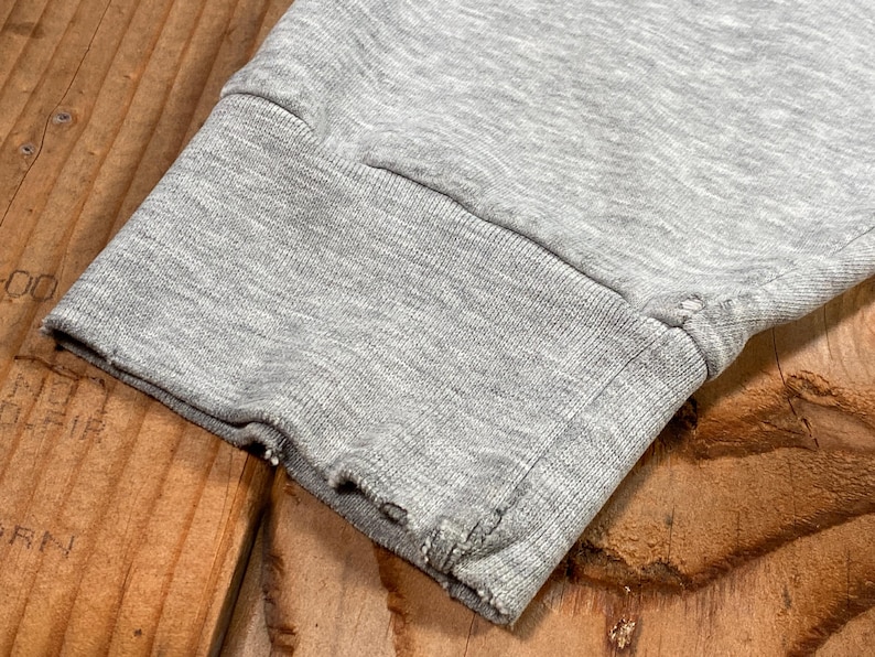 Puede incluir: Primer plano del pu&ntilde;o de una manga de sudadera gris. El pu&ntilde;o es acanalado y tiene un borde deshilachado y desgastado. La tela es gris jaspeado y la manga descansa sobre una superficie de madera.