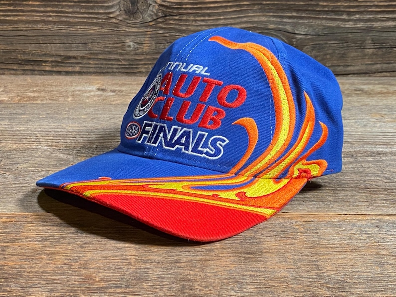 Puede incluir: Gorra de b&eacute;isbol azul con el texto "Annual Auto Club Finals" en rojo y blanco. La gorra presenta un dise&ntilde;o de llama en amarillo y naranja, con una visera roja.