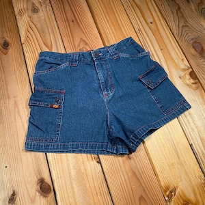 May include: Blue denim shorts with cargo pockets and red stitching. The shorts have a button and zipper closure and are made of a sturdy denim material. The shorts have the brand name "ROXY" on the cargo pocket.