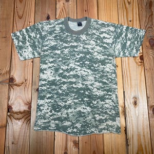 May include: A short-sleeved t-shirt with a digital camouflage pattern in shades of green, gray, and white. The shirt has a crew neck and a tag at the neckline. The fabric appears to be a lightweight material.