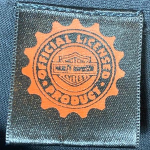 May include: A black fabric label with an orange gear-shaped border. The label reads "OFFICIAL LICENSED PRODUCT" around the edge, with "MOTOR HARLEY-DAVIDSON CYCLES" in the center. The label is rectangular.