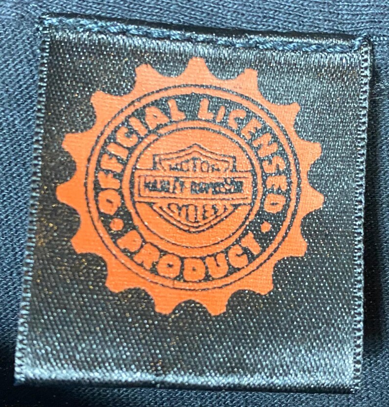 May include: A black fabric label with an orange gear-shaped border. The label reads "OFFICIAL LICENSED PRODUCT" around the edge, with "MOTOR HARLEY-DAVIDSON CYCLES" in the center. The label is rectangular.