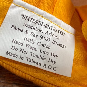 May include: Close-up of a white tag with black text, stitched to a yellow fabric. The tag reads "STATESIDE-ENTERTEC" and includes care instructions: "Hand Wash. Line Dry. Do Not Tumble Dry." The fabric is 100% cotton.