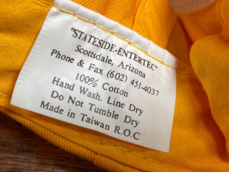 May include: Close-up of a white tag with black text, stitched to a yellow fabric. The tag reads "STATESIDE-ENTERTEC" and includes care instructions: "Hand Wash. Line Dry. Do Not Tumble Dry." The fabric is 100% cotton.