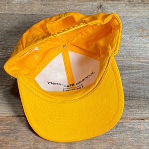 May include: A bright yellow baseball cap with a curved brim and adjustable snapback closure. The interior features a white lining with black text. The cap is made of a durable material and is designed for sun protection.