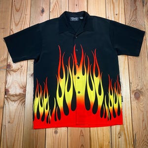 May include: Black short-sleeved button-up shirt with a flame design in red, yellow, and orange. The flames start at the bottom and extend upwards. The shirt has a collar and a few buttons down the front.