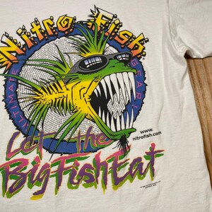 Puede incluir: Camiseta blanca con un gr&aacute;fico colorido de un pez con gafas de sol. El dise&ntilde;o incluye el texto "Nitro Fish Ultimate Gear" y la frase "Let the Big Fish Eat."