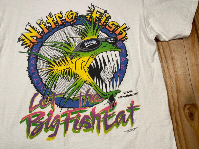 Puede incluir: Camiseta blanca con un gr&aacute;fico colorido de un pez con gafas de sol. El dise&ntilde;o incluye el texto "Nitro Fish Ultimate Gear" y la frase "Let the Big Fish Eat."