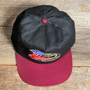 May include: A black baseball cap with a maroon peak. The cap has a small, embroidered patch with a design incorporating red, white, and blue elements. The cap has a maroon button on top.