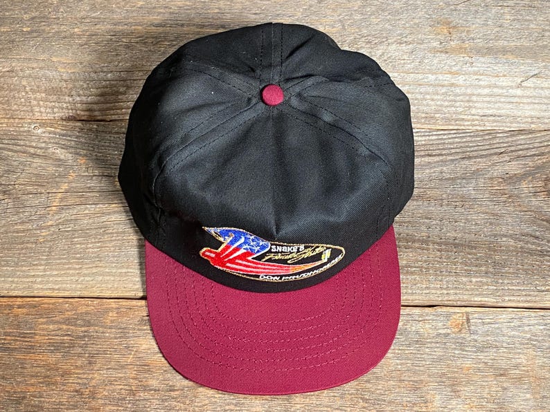 May include: A black baseball cap with a maroon peak. The cap has a small, embroidered patch with a design incorporating red, white, and blue elements. The cap has a maroon button on top.