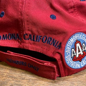 May include: A red baseball cap featuring dark blue embroidered text that reads POMONA, CALIFORNIA and MEMBERS ONLY. A circular patch on the side displays the AAA logo with the text AUTOMOBILE CLUB OF SOUTHERN CALIFORNIA in blue and white.