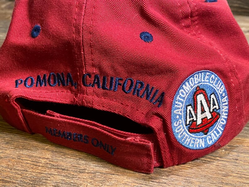 May include: A red baseball cap featuring dark blue embroidered text that reads POMONA, CALIFORNIA and MEMBERS ONLY. A circular patch on the side displays the AAA logo with the text AUTOMOBILE CLUB OF SOUTHERN CALIFORNIA in blue and white.