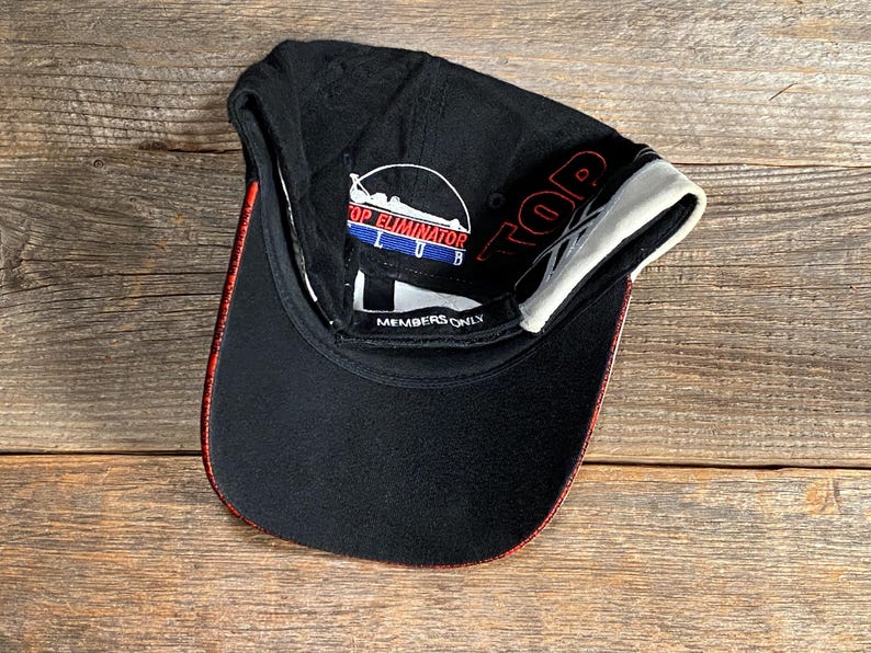 May include: Black baseball cap with red and white accents. The cap features the text "TOP ELIMINATOR CLUB" and "MEMBERS ONLY". The brim has a red trim and the interior is white.
