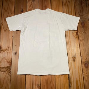 May include: A plain white t-shirt with short sleeves, laid flat on a wooden surface. The shirt is centered in the frame, with the back side facing up. The fabric appears to be cotton or a similar material.