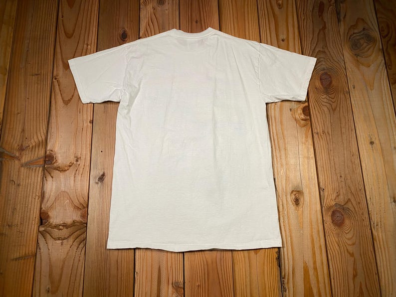 May include: A plain white t-shirt with short sleeves, laid flat on a wooden surface. The shirt is centered in the frame, with the back side facing up. The fabric appears to be cotton or a similar material.