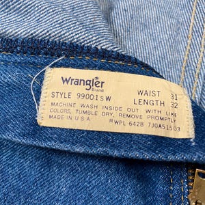 May include: Close-up of blue denim jeans with a Wrangler brand tag. The tag indicates a waist size of 31 inches and a length of 32 inches. The tag also includes washing instructions and the text "MADE IN USA".