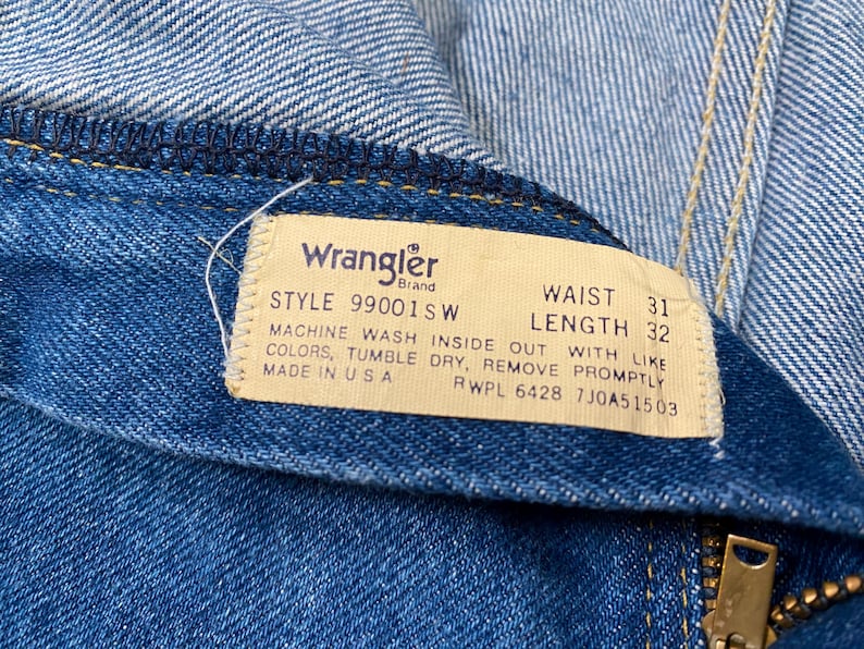 May include: Close-up of blue denim jeans with a Wrangler brand tag. The tag indicates a waist size of 31 inches and a length of 32 inches. The tag also includes washing instructions and the text "MADE IN USA".