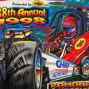 May include: White t-shirt featuring a colorful graphic of a drag racing car. The design includes the text "38th Annual 1998" and logos for Pennzoil, NHRA, and Chief Auto Parts. The car is red and blue with a driver wearing a red helmet.