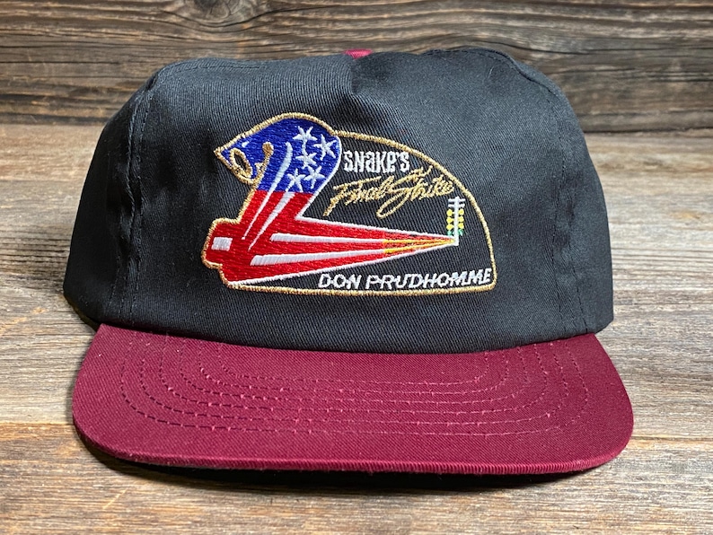 May include: Black and burgundy baseball cap with a flat brim. The cap features an embroidered patch with the text "Snake's Final Strike" and a design incorporating the American flag. The text "DON PRUDHOMME" is also visible.