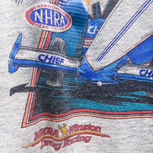 May include: A gray t-shirt with a colorful graphic of a drag racing car. The graphic includes the text "Chief" and "NHRA Championship Drag Racing". The bottom of the graphic includes the text "NHRA Winston Drag Racing".