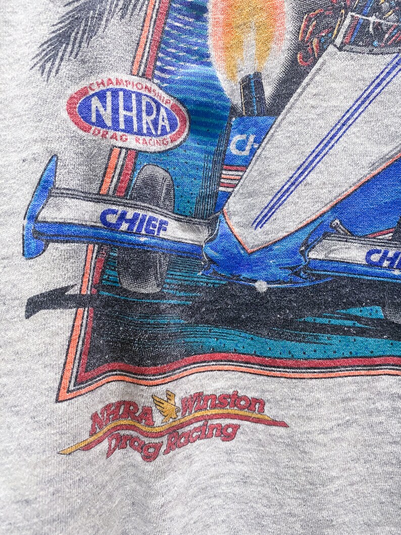 May include: A gray t-shirt with a colorful graphic of a drag racing car. The graphic includes the text "Chief" and "NHRA Championship Drag Racing". The bottom of the graphic includes the text "NHRA Winston Drag Racing".