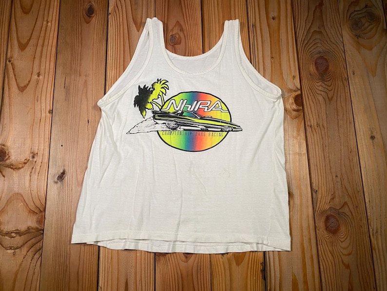 Pu&ograve; includere: Canotta bianca con una grafica colorata di un'auto da drag racing, palme e la scritta "NHRA Championship Drag Racing" in un cerchio arcobaleno. La canotta &egrave; realizzata in un materiale morbido e leggero.