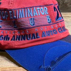 May include: A red baseball cap with a blue brim featuring embroidered text that reads TOP ELIMINATOR CLUB 21, NATIONAL HOT ROD, and 36th ANNUAL AUTO CLUB. The blue brim includes a stitched graphic of a race car silhouette under a circular arc.