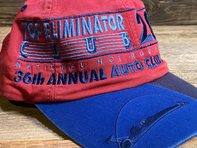 May include: A red baseball cap with a blue brim featuring embroidered text that reads TOP ELIMINATOR CLUB 21, NATIONAL HOT ROD, and 36th ANNUAL AUTO CLUB. The blue brim includes a stitched graphic of a race car silhouette under a circular arc.