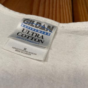 May include: Close-up of a white Gildan Activewear Ultra Cotton t-shirt. The tag reads "Gildan Activewear Ultra Cotton Heavyweight" with the size "M" and "Made in Canada". The shirt is made of 100% pre-shrunk cotton.