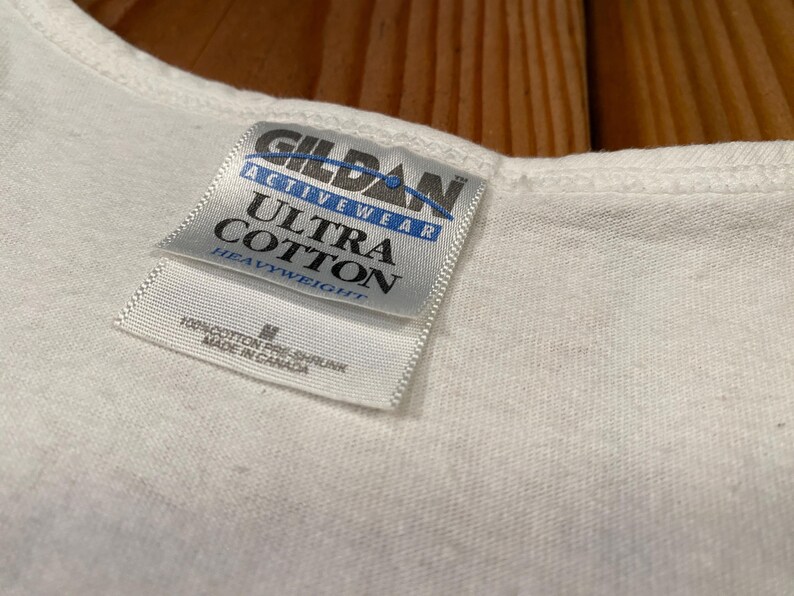 May include: Close-up of a white Gildan Activewear Ultra Cotton t-shirt. The tag reads "Gildan Activewear Ultra Cotton Heavyweight" with the size "M" and "Made in Canada". The shirt is made of 100% pre-shrunk cotton.