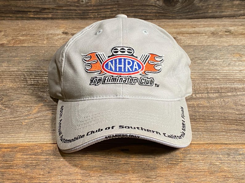 May include: A light grey baseball cap with the NHRA Top Eliminator Club logo in orange, white, and blue. The cap also displays text that reads "40th Annual Automobile Club of Southern California NHRA Finals".