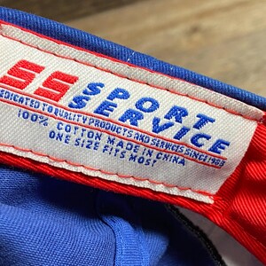 Puede incluir: Primer plano de una gorra de b&eacute;isbol azul con borde rojo. La etiqueta interior dice "SS SPORT SERVICE" con texto que indica que la gorra es 100% algod&oacute;n y talla &uacute;nica.